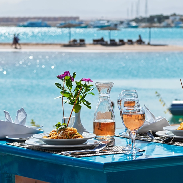 Table set overlooking a marina, featuring a delicious pasta dish and a glass of wine. The table is elegantly arranged with tableware and a view of the boats and water in the background, creating a sophisticated and relaxing dining experience.