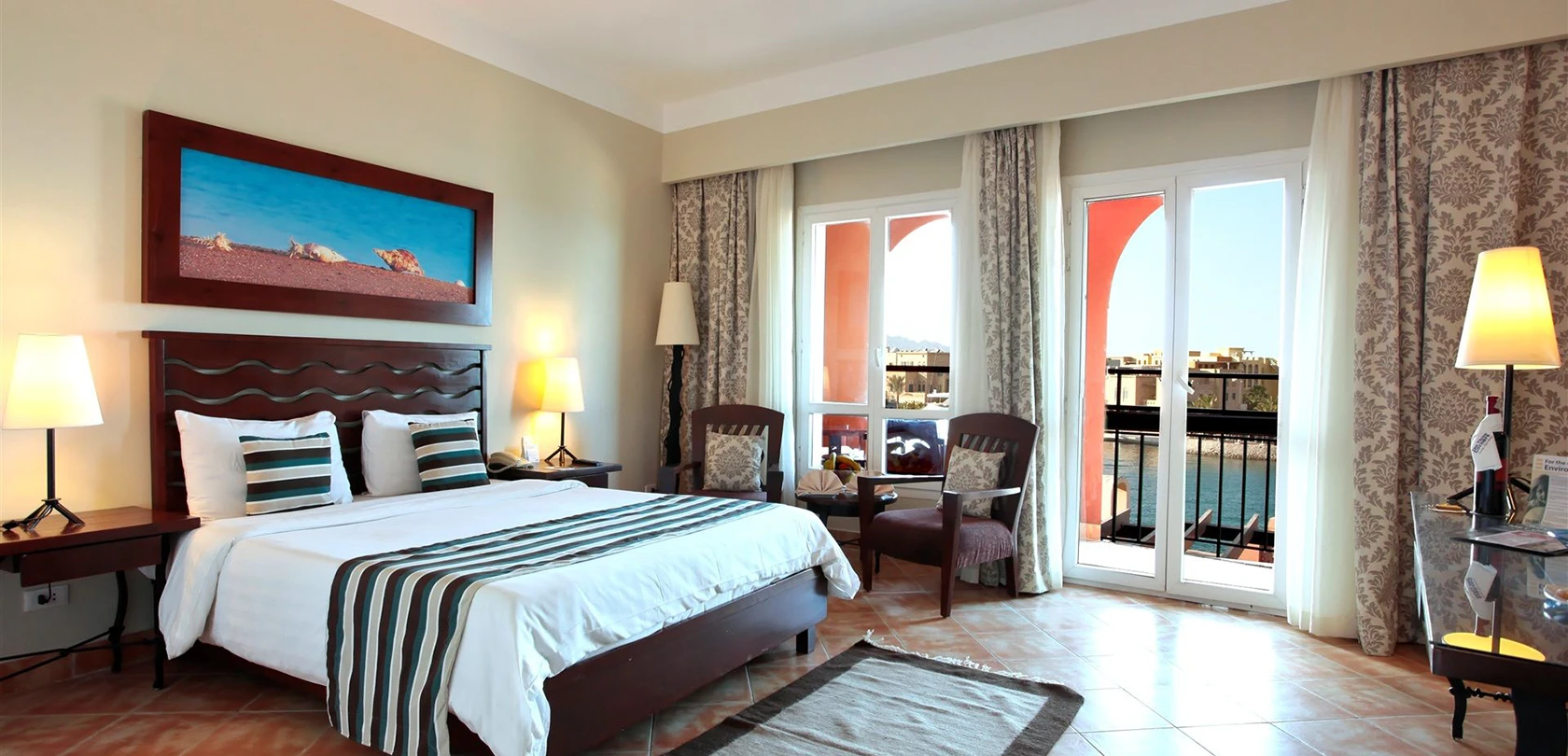 Ocean view standard room at Three Corners - Ocean View, featuring an open curtain balcony with a breathtaking view of the sea. The room includes a large bed, two chairs, and a painting on the wall, creating a cozy and artistic atmosphere.