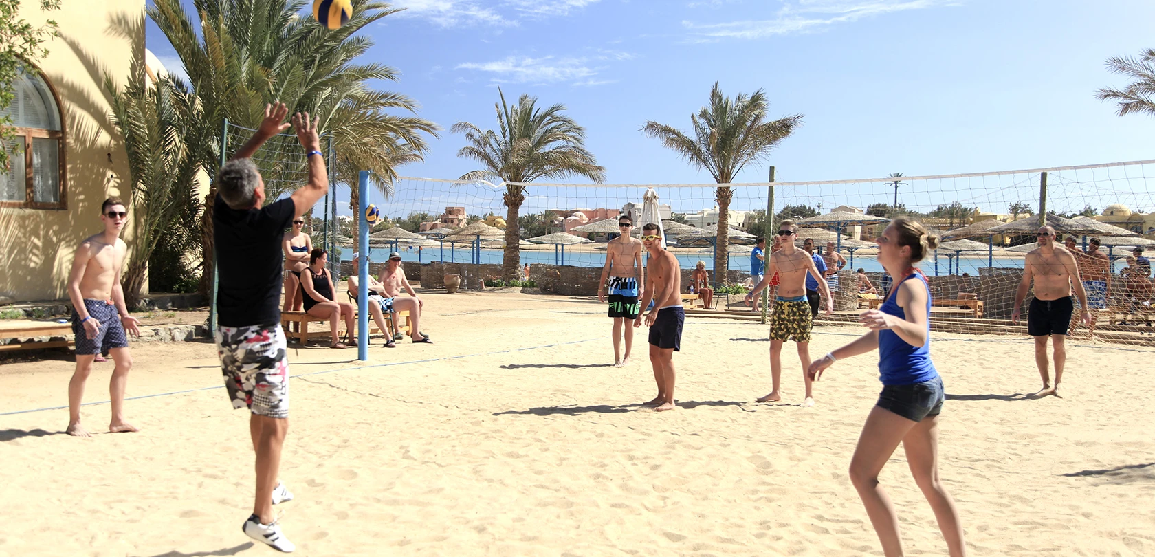 People playing beach volleyball near the shore, with the ocean and sandy beach as the backdrop. The scene captures an active and enjoyable beach activity in a sunny, outdoor setting.
