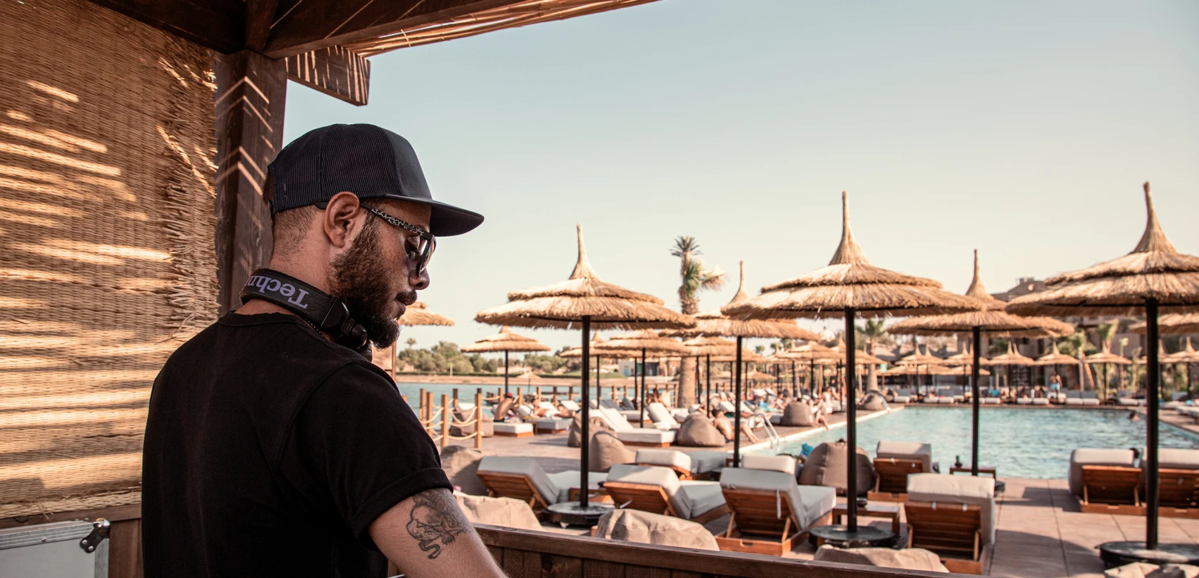 DJ performing by the pool at Cooks Club El Gouna, setting a lively and energetic atmosphere. The DJ booth is positioned near the poolside, with speakers and equipment arranged for optimal sound. Guests can be seen relaxing on lounge chairs or swimming, enjoying the vibrant music. The scene is enhanced by sunny weather and clear blue skies, creating a perfect backdrop for a fun and social poolside experience.