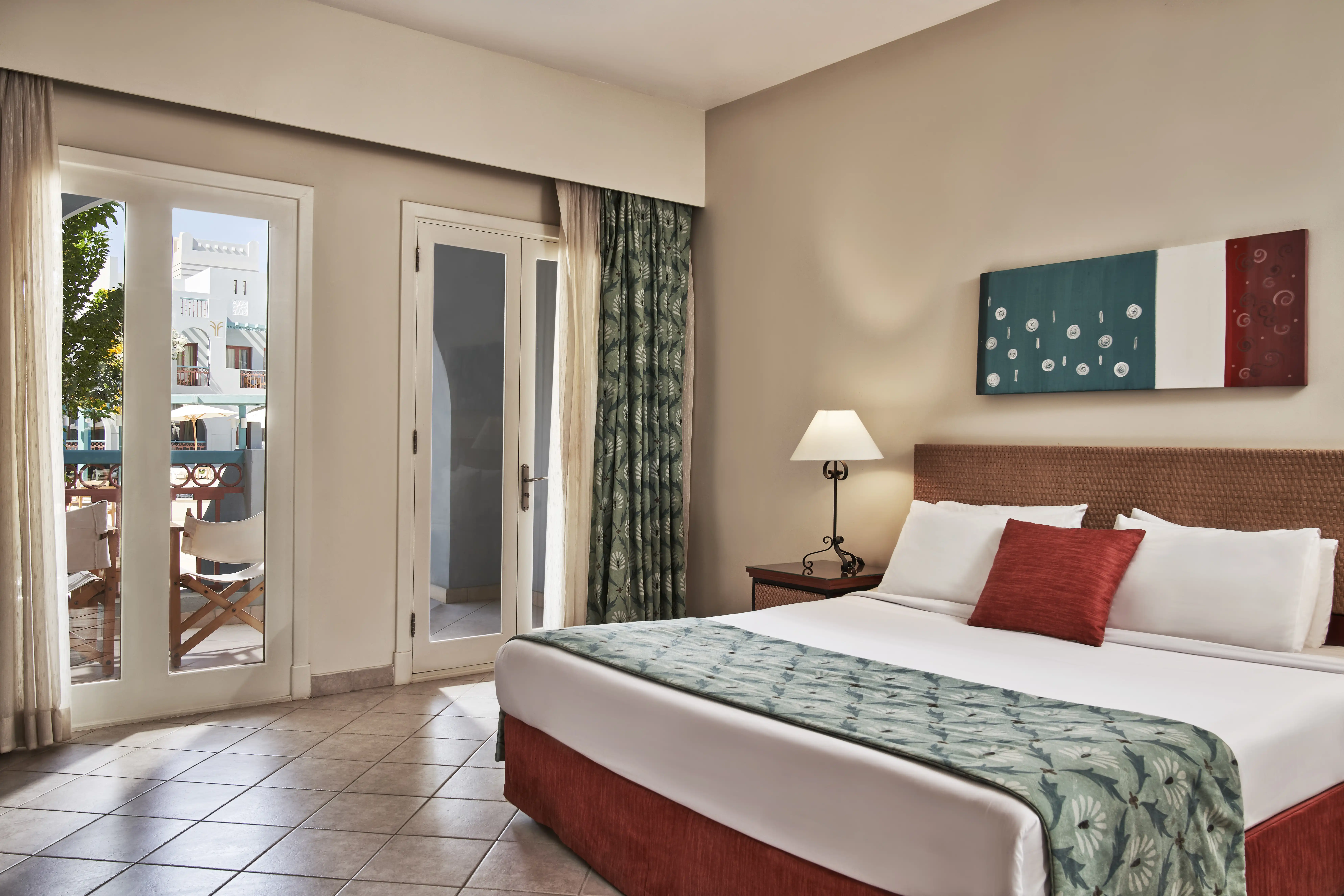 Spacious bedroom with a large bed in a green-blueish room, featuring an open balcony with a seating area that includes two chairs on the terrace, offering a serene and inviting atmosphere.