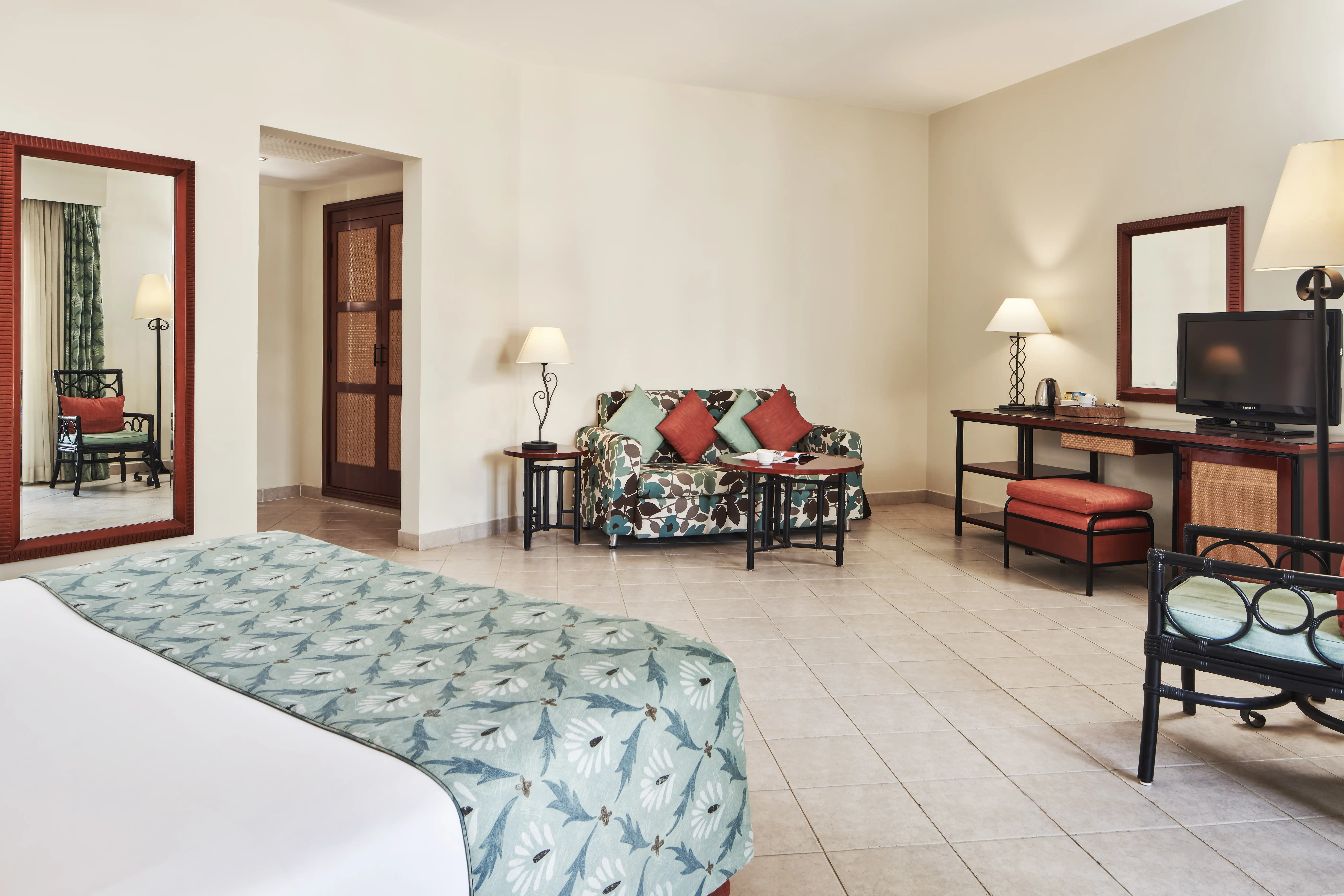 Spacious bedroom with a large bed in a green-blueish room, featuring a television, a seating area with a sofa, a chair, and a table, along with a mirror table adding elegance to the space.